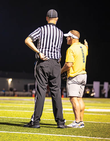 Two adults stand on a football field at night, one wearing a black and white striped referee shirt and the other wearing a yellow shirt with “WILDCATS" written on the back, engaged in conversation. Description generated by AI
