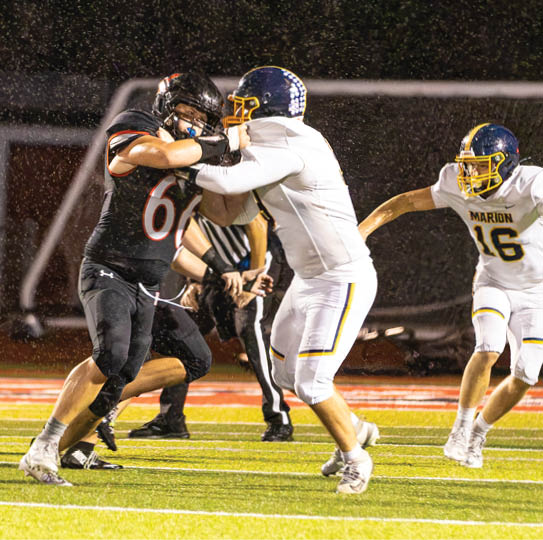 Two people in football uniforms are engaged in a physical block on a rain-soaked field while a third player in a white uniform runs toward them. Description generated by AI