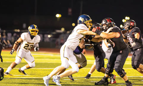 Two football players in white uniforms with blue and yellow helmets are engaged in blocking against players in black and red uniforms on a lit field at night. Description generated by AI