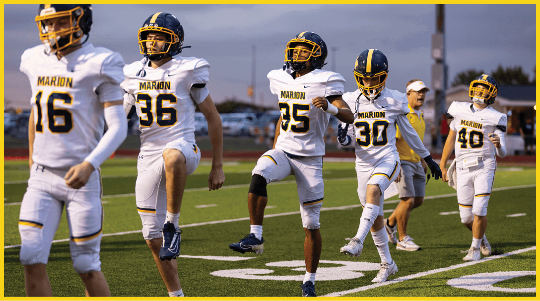 Three people in white football uniforms with the word Marion on the jerseys are on a field, with one person raising a knee as if warming up or stretching. Description generated by AI