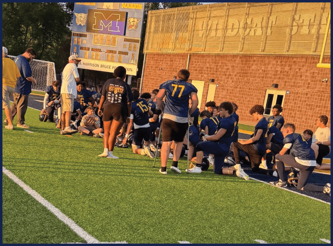 A group of people in sports uniforms and casual clothing are gathered on a field near a scoreboard during what appears to be a team meeting or practice. Description generated by AI