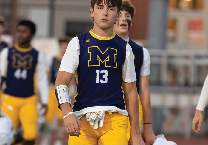 Two adults wearing yellow football pants and navy blue jerseys with a large yellow M are walking on a field carrying white helmets. Description generated by AI