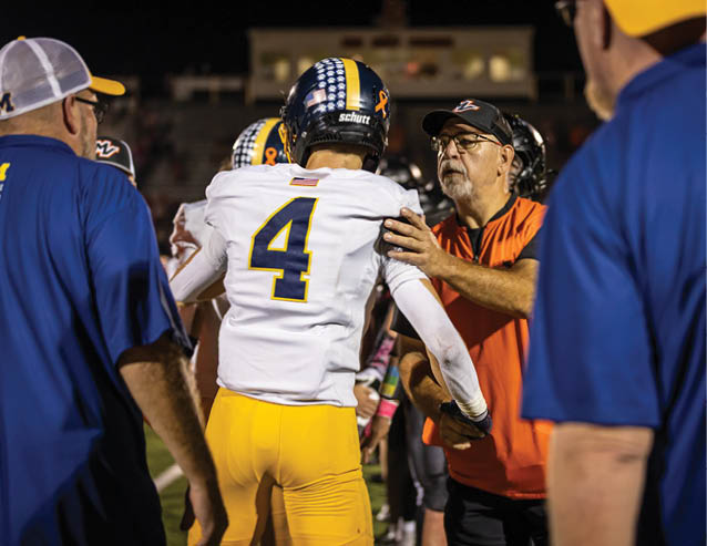 A person wearing a football uniform with the number 4 is being patted on the shoulder by another person in an orange shirt and cap on a sports field at night. Description generated by AI