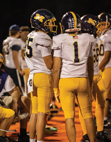 Two people wearing football helmets and white jerseys are standing close together, with one jersey displaying the number one and American flag patches. Description generated by AI