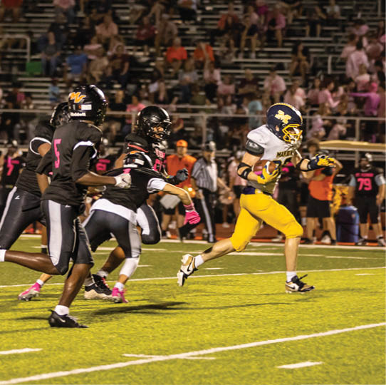 A group of people in football uniforms are actively running on a field during a game, with one person in yellow pants holding the ball and others in black uniforms chasing. Description generated by AI