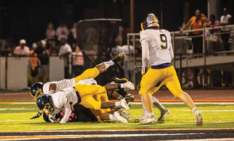 Several people in football uniforms are engaged in a tackle on a brightly lit field during a nighttime game. Description generated by AI