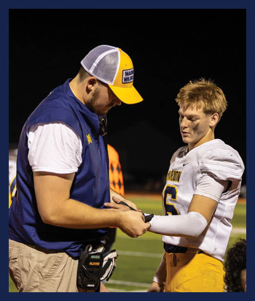 An adult wearing a yellow and white cap and a blue vest is holding the wrist of a person in a white football uniform with the number six on it during a nighttime outdoor event. Description generated by AI