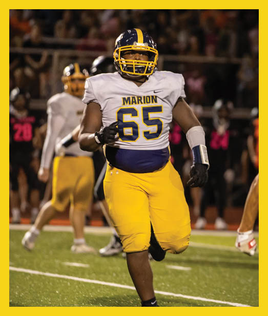 A person wearing a Marion football uniform with the number 65 is running on a football field during a game. Description generated by AI