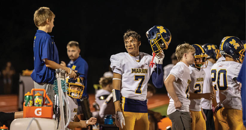 A group of children in football uniforms and casual clothing are gathered on a sports field at night, with one child holding a helmet and others standing or sitting nearby. Description generated by AI