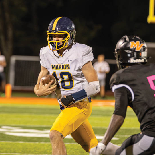 A person wearing a football uniform with a helmet and holding a football is running on a field during a nighttime game. Description generated by AI