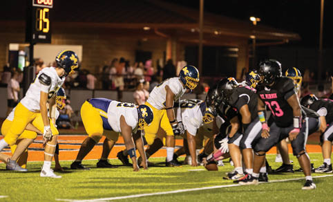 Two opposing football teams in yellow and black uniforms are lined up on the field preparing for a play under stadium lights at night. Description generated by AI