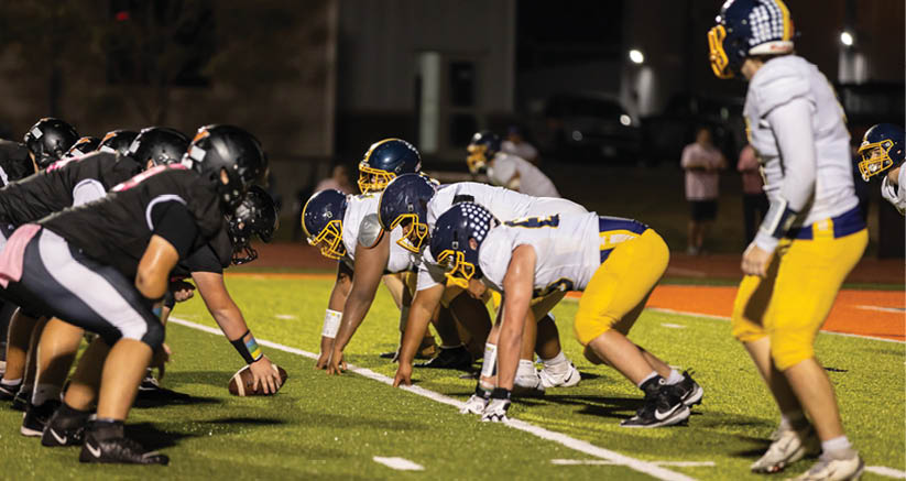 Two opposing football teams in helmets and uniforms are lined up facing each other on a field under stadium lights during a night game. Description generated by AI
