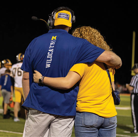 Two people wearing sports apparel with one person wearing a headset and a cap labeled Marion Wildcats are embracing on a sports field at night. Description generated by AI