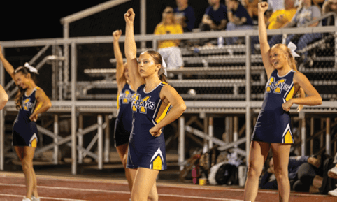 Adults in matching athletic uniforms with the word Wildcats perform a synchronized cheer routine with raised fists in front of bleachers. Description generated by AI