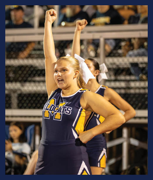An adult in a cheerleading uniform with a Wildcats logo is raising one arm enthusiastically while standing in front of another person in a similar outfit at a sports event. Description generated by AI