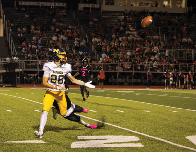 A person in a yellow and white football uniform is throwing a football while another person in a dark uniform attempts to tackle them on a lit football field with spectators in the background. Description generated by AI
