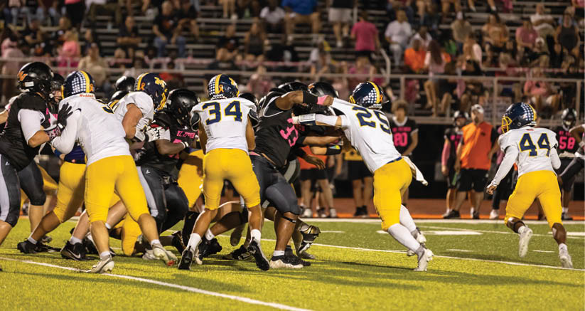 People in football uniforms are actively engaged in a play on a brightly lit field with a crowd watching from the stands. Description generated by AI