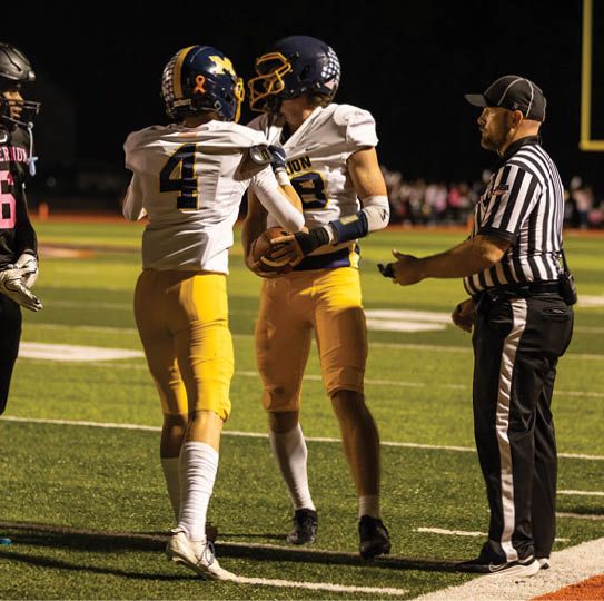 A person wearing a referee uniform is holding a whistle and standing next to a person in a football uniform on a sports field at night. Description generated by AI