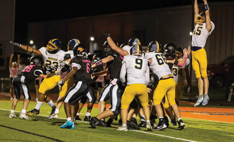 A group of people wearing football uniforms are engaged in a play on a field at night with one person jumping to catch or block the ball. Description generated by AI