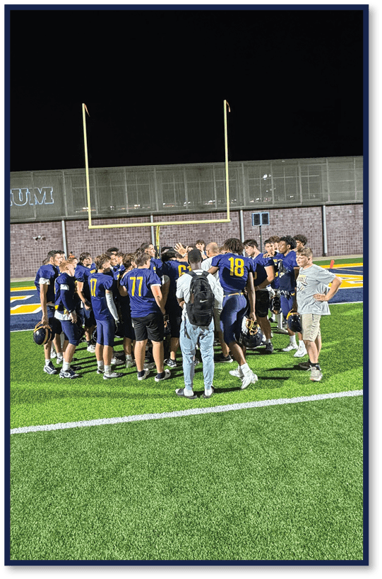 A group of people wearing dark sports uniforms with numbers are gathered on a football field near the goalpost at night. Description generated by AI
