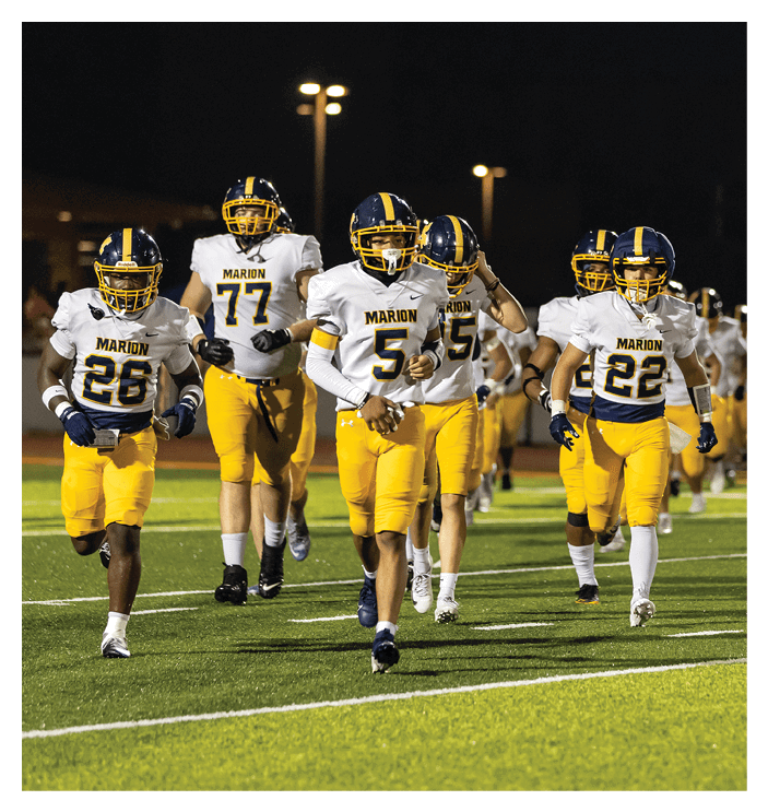 A group of people wearing yellow pants and white football jerseys with helmets are running on a football field at night. Description generated by AI