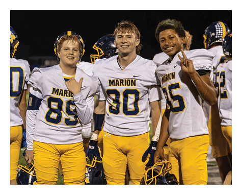 Three people wearing white football jerseys with “Marion" written on them smile and pose together at night, with two making hand gestures. Description generated by AI