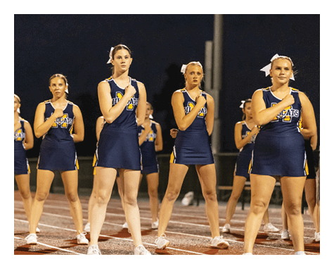 A group of adults in matching cheerleading uniforms stand in formation on a track at night with one hand placed over their chests. Description generated by AI