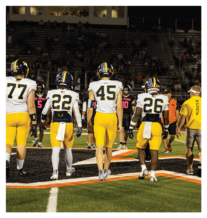 Four people in yellow and white football uniforms walk on a field toward a group of people in black and pink uniforms. Description generated by AI