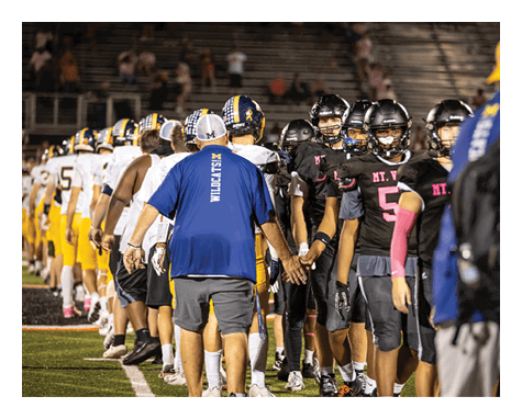 People in football uniforms and coaches in team shirts are shaking hands on a field with empty bleachers in the background. Description generated by AI