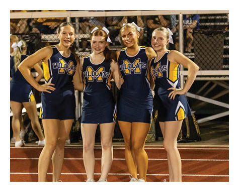 Four adults wearing matching Wildcats cheerleading uniforms stand side by side on a track at night with a fence and bleachers in the background. Description generated by AI