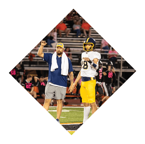 Two people stand on a football field holding hands, one wearing yellow pants and the other wearing gray shorts. Description generated by AI