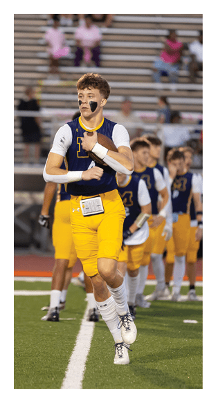 A person wearing a sports jersey with face paint is holding a football on a field with blurred spectators in the background. Description generated by AI