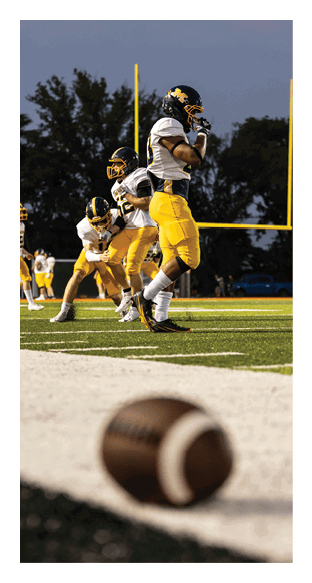 Several people in yellow and white football uniforms are on a field near a goalpost with a football lying on the ground in the foreground. Description generated by AI