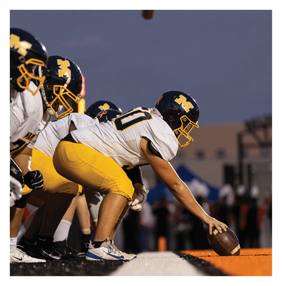 Several people wearing dark helmets with a yellow “M" logo and white sports uniforms are positioned under a football goalpost against a clear sky. Description generated by AI