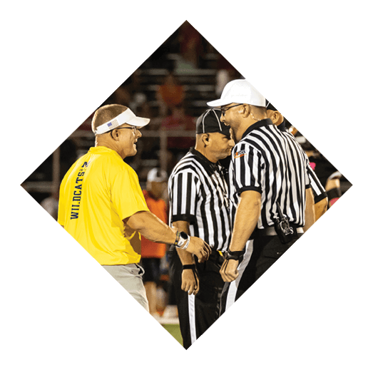 Three adults are engaged in a conversation on a sports field, with two wearing black and white striped referee uniforms and one wearing a yellow shirt with “WILDCATS" written on the back. Description generated by AI