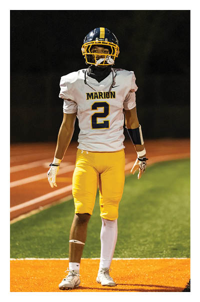 A person wearing a football uniform with a helmet, white jersey with the word “MARION" and number 2, yellow pants, and white cleats is standing on an orange and green football field. Description generated by AI
