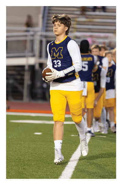 A person wearing a blue and yellow sports uniform with the number 33 is holding a football and walking on a field with other similarly dressed people in the background. Description generated by AI