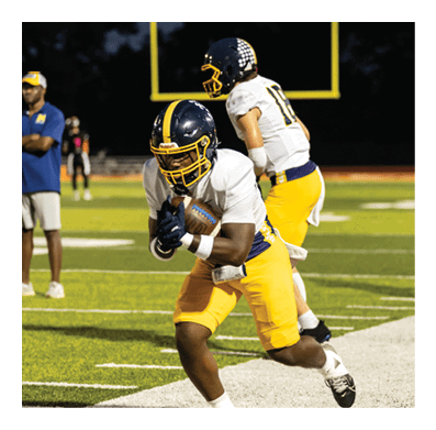 Two people wearing football helmets and uniforms are on a field with a goalpost visible in the background during a nighttime game. Description generated by AI