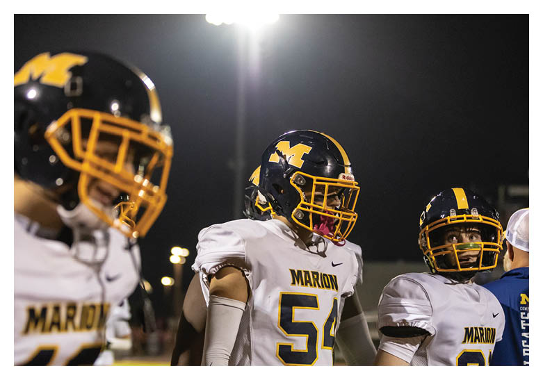 Three people wearing football helmets and white jerseys with “MARION" written on them stand on a field under bright stadium lights at night. Description generated by AI