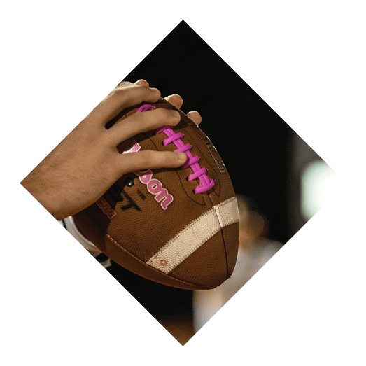 A person is holding a football with pink laces on a field with blurred players and a scoreboard in the background. Description generated by AI