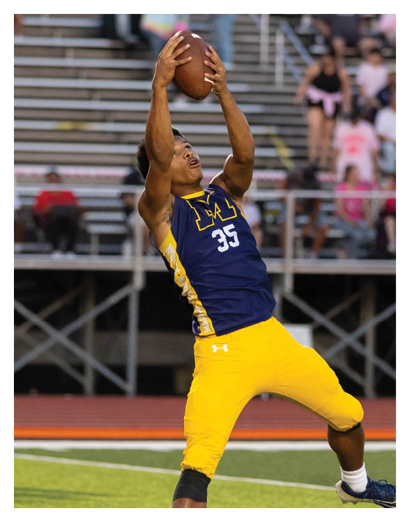 A person wearing a blue and yellow sports uniform is reaching up to catch a football on a field with bleachers and spectators in the background. Description generated by AI