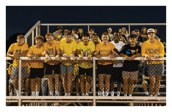 A group of smiling young people wearing casual clothing, including yellow and white shirts, are standing closely together behind a railing at an outdoor event with a crowd in the background. Description generated by AI