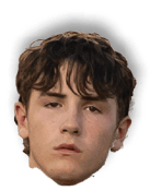 A person with short, wavy hair looks directly at the camera with a neutral expression, while another person is partially visible in the background. Description generated by AI