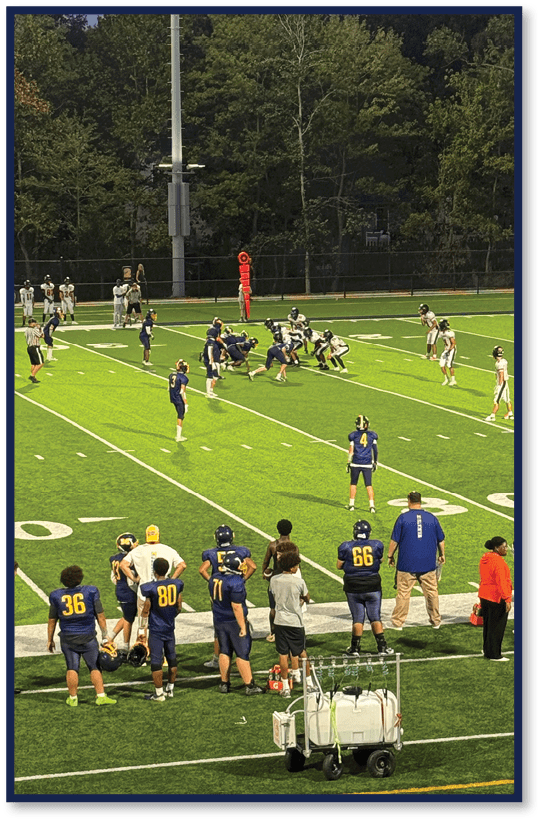 A group of people in sports uniforms are on a football field with some standing on the sidelines and others positioned on the field near the line of scrimmage. Description generated by AI