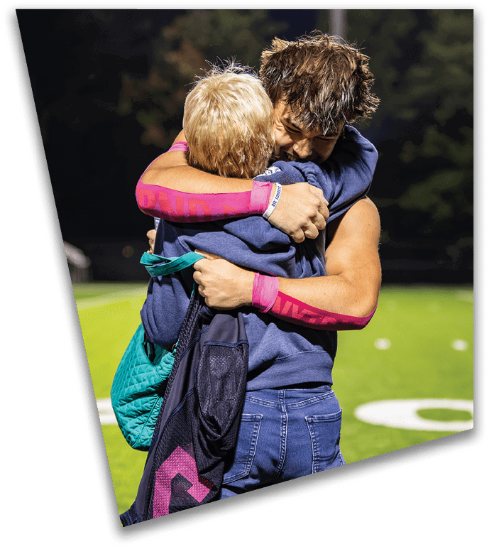 Two people wearing casual clothing embrace tightly on a sports field at night with trees and a light pole in the background. Description generated by AI
