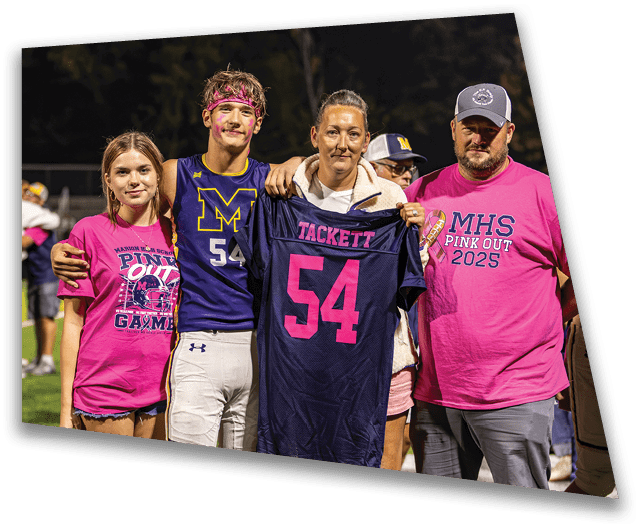 Four people stand together outdoors at night with one person holding a sports jersey while two others wear pink shirts supporting a 2025 event. Description generated by AI
