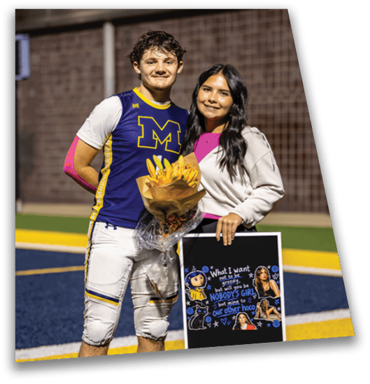 Two adults stand on a sports field with one holding a bouquet of flowers and the other holding a decorated poster. Description generated by AI
