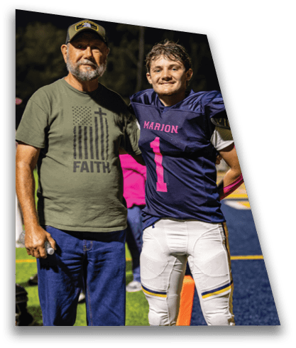 An adult wearing a green shirt with the word faith and a cap stands next to a person in a blue and pink football uniform with the number one on a sports field at night. Description generated by AI