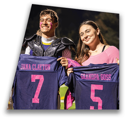 Two people smiling and holding up sports jerseys with names and numbers on them, one wearing shoulder pads and face paint, standing outdoors at night. Description generated by AI