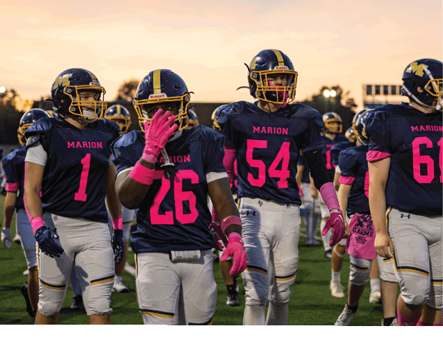 A group of people wearing football uniforms with helmets and pink gloves are walking together on a field during sunset. Description generated by AI